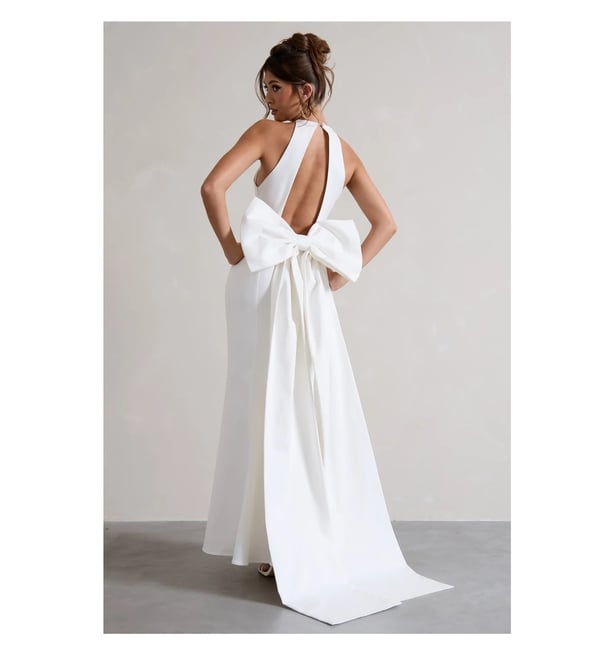 Model wears white open back sleeveless halterneck dress with back bow detailing